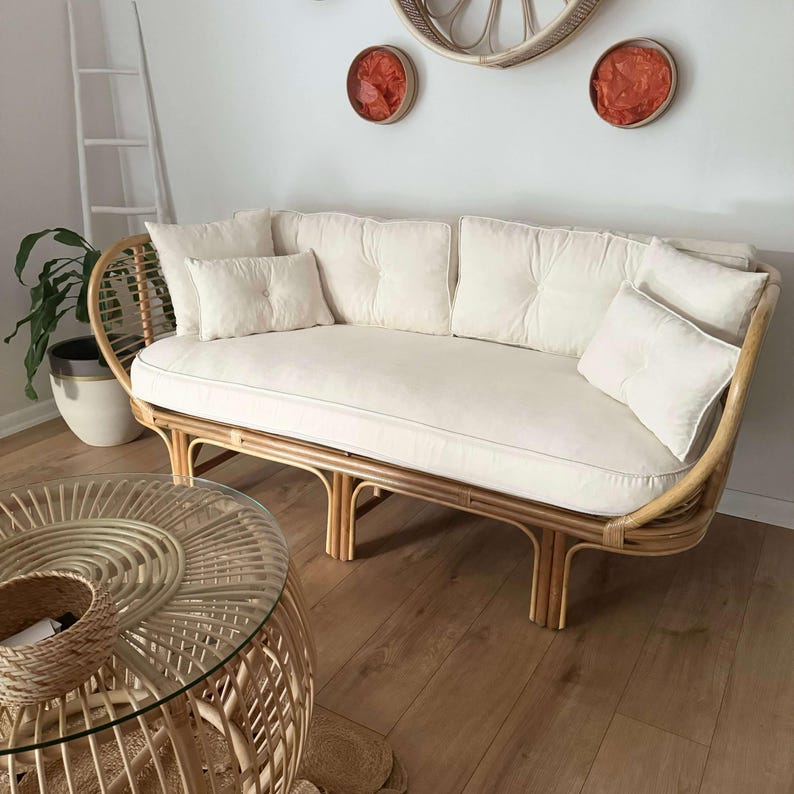 May include: A white rattan sofa with a white cushion and pillows. The sofa has a curved back and is sitting on a wooden floor.