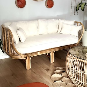 May include: A light brown rattan sofa with a white cushion and three pillows. The sofa has a curved back and a low profile. The sofa is sitting on a wooden floor.