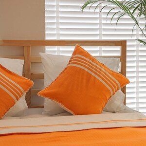 May include: Two white pillowcases with orange and white striped throw pillows on a bed with a white and orange striped blanket.