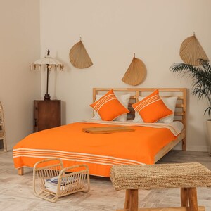 May include: A bright orange bed with white stripes and a wooden frame. The bed has two orange pillows and a wooden tray on top of the bedding. The bed is in a room with white walls and a wooden side table.