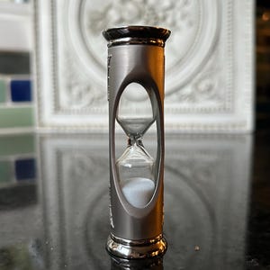 May include: A silver hourglass with a black top and base. The hourglass has a clear glass center filled with white sand. The hourglass is standing on a black surface.