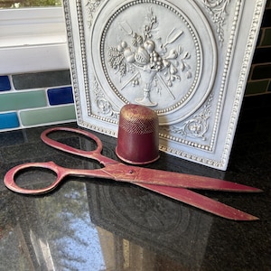 Könnte beinhalten: Eine Schere aus rot-goldfarbenem Metall mit Used-Look neben einem passenden Fingerhut in Rot und Gold. Dahinter steht eine rechteckige, hellgraue Zierplatte mit einem zentralen Relief, das einen Obstkorb darstellt.