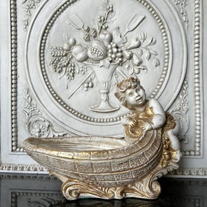 Puede incluir: Centro de mesa decorativo, en tonos blanco roto y dorado, con temática de querubines. El centro de mesa presenta un cuenco en forma de barco con una figura de querubín en el lateral. El fondo es un relieve blanco con un diseño floral y frutal.