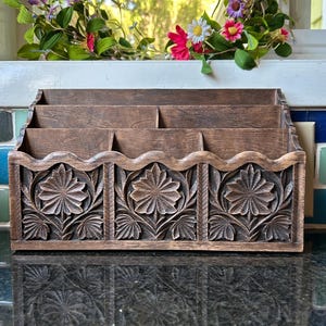 Peut inclure: Un organisateur de bureau en bois brun foncé doté de trois compartiments étagés pour le rangement. Le panneau avant est orné de trois motifs floraux sculptés, chacun encadré dans une section rectangulaire, surmonté d'un bord ondulé.