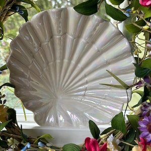 May include: A scallop-shaped ceramic dish with a glossy, iridescent finish that reflects light in soft pastel tones. The dish features deep, radiating ridges that mimic the natural texture of a seashell, creating a fan-like pattern.
