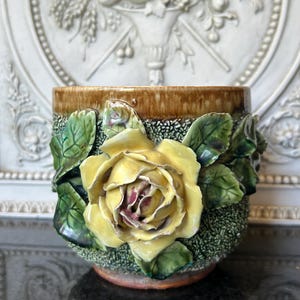 Puede incluir: Jardinera de cerámica con una rosa amarilla y hojas verdes. La jardinera tiene una base verde texturizada y un borde marrón. La rosa es el punto focal, con pétalos detallados y un toque de rojo en el centro. Diseño floral decorativo.