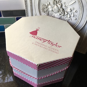 Könnte beinhalten: Weiße achteckige Schachtel mit rosa und weißen Streifen. Die Schachtel hat ein rotes Logo mit der Aufschrift "Nancy Taylor Finishing School & Modeling Studio".