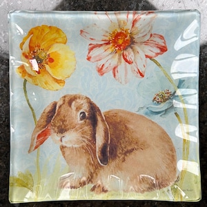 Puede incluir: Plato decorativo cuadrado de vidrio con un conejo marrón de orejas largas, rodeado de flores amarillas y rojas. El fondo es azul claro con un patrón sutil. El plato tiene un borde transparente ligeramente elevado.