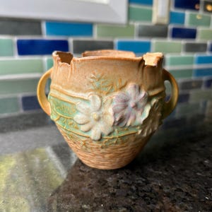 Peut inclure: Un récipient en céramique avec deux anses latérales, doté d'une base texturée beige et d'une bande de motifs floraux sculptés en relief dans des tons vert pâle, rose tendre et crème. Le bord est festonné et irrégulier, avec une finition mate.