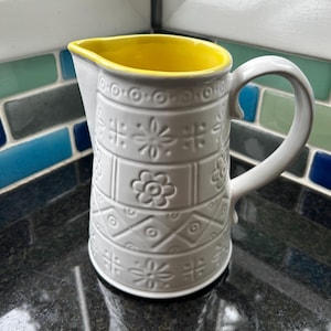 May include: White ceramic pitcher with a yellow rim and a geometric pattern. The pitcher has a handle and a wide mouth.