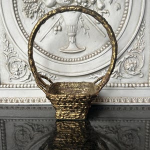 May include: A gold-colored decorative basket with a woven texture and a rounded handle. The basket is set against a white background with a floral relief design. The basket's handle is approximately 8 inches in height.