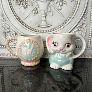 Pode incluir: Duas canecas de Páscoa de cerâmica. Uma caneca apresenta um design de coelho com uma borda rosa e amarela. A outra caneca tem a forma de um coelho usando macacão azul. Ambas as canecas têm alças e são brancas.