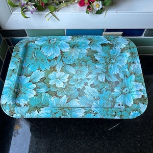 May include: A rectangular metal serving tray featuring a vibrant floral pattern in shades of bright turquoise, light blue, and dark green. The tray has rounded corners, raised edges, and a glossy finish with visible metallic rivets at the corners.
