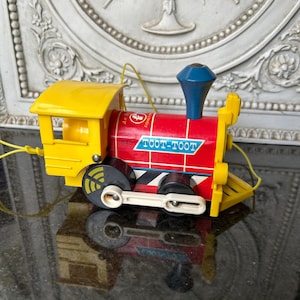 May include: A vintage toy train with a yellow cab and red body. The train has black wheels, a blue smokestack, and the words "TOOT-TOOT" printed on the side. A yellow string is attached for pulling.