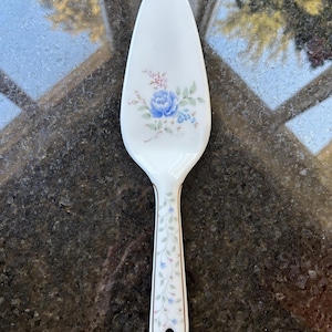 Puede incluir: Un servidor de pastel de cerámica blanca con un diseño floral con flores azules y rosas. El mango está decorado con un patrón de vid.