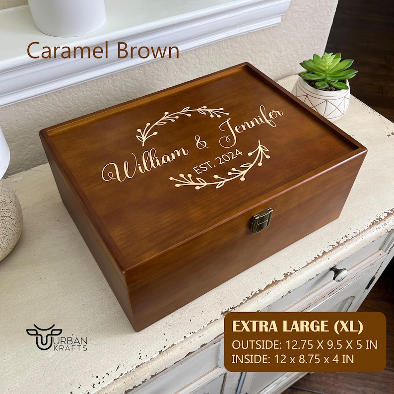 Pu&ograve; includere: Una scatola di legno extra large, marrone caramello, con coperchio incernierato. Il coperchio &egrave; inciso con "William & Jennifer" e "EST. 2024" in oro, incorniciato da elementi floreali. Dimensioni esterne: 32.39 x 24.13 x 12.7 cm, interne: 30.48 x 22.23 x 10.16 cm.