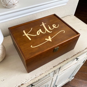 May include: A rectangular wooden box with a hinged lid. The box is stained a warm brown color and features the name "Katie" in a gold script font on the lid, along with a heart symbol. A metal clasp secures the box.