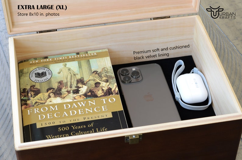 Pu&ograve; includere: Scatola di legno aperta con esterno marrone scuro e interno in legno chiaro. All'interno, un libro intitolato "From Dawn to Decadence" e uno smartphone poggiano su una fodera in velluto nero. &Egrave; visibile anche una custodia bianca per auricolari wireless.