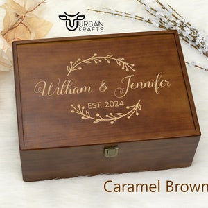 Pu&ograve; includere: Una scatola di legno marrone caramello con coperchio incernierato. La scatola &egrave; incisa con i nomi "William & Jennifer" e l'anno "EST. 2024" all'interno di una ghirlanda floreale decorativa. La scatola ha una chiusura in metallo ed &egrave; su sfondo bianco.