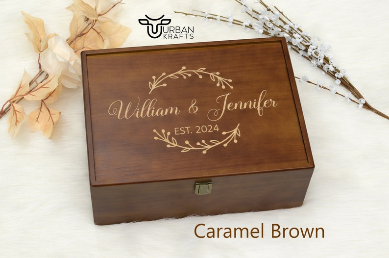 Pu&ograve; includere: Una scatola di legno marrone caramello con coperchio incernierato. La scatola &egrave; incisa con i nomi "William & Jennifer" e l'anno "EST. 2024" all'interno di una ghirlanda floreale decorativa. La scatola ha una chiusura in metallo ed &egrave; su sfondo bianco.