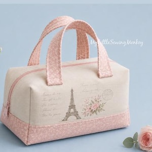 May include: A rectangular fabric handbag with two top handles, featuring a beige main body with a printed Eiffel Tower, floral bouquet, and script text. The base and handles are made of pink fabric with a small white floral pattern. A pink zipper.