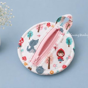 May include: A round fabric coin purse featuring a white background with a repeating pattern of Little Red Riding Hood, wolves, trees, and mushrooms. The pouch has a pink zipper closure across the center and a small fabric loop at the top.