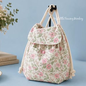 May include: A drawstring backpack made of cream-colored fabric with a repeating floral pattern of pink flowers and green leaves. The bag features a front pocket with a single button closure, a fold-over flap, and thick white rope shoulder straps.