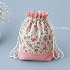 May include: A drawstring bag featuring a cream-colored fabric with a multi-colored floral print, a solid pink base, and a pink ruffled top edge. The bag includes white cord straps and the text MyLittleSewingMonkey printed on the front surface.