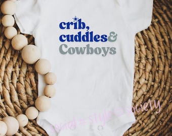 May include: White baby onesie with the text "crib, cuddles & Cowboys" in blue and gray. The onesie is on a woven basket with wooden beads.