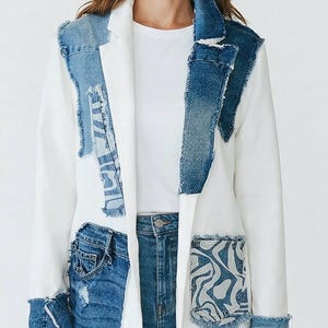 May include: White blazer with denim patchwork details, including the collar, pockets, and cuffs. The jacket features a classic lapel and a tailored fit. The model is wearing a white t-shirt and blue jeans.