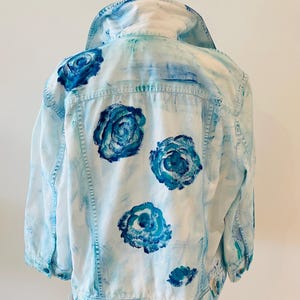 May include: A light blue denim jacket with hand-painted blue rose designs on the back. The jacket has a classic collar, button closures, and pockets. The roses are a focal point, adding a unique artistic touch to the garment.