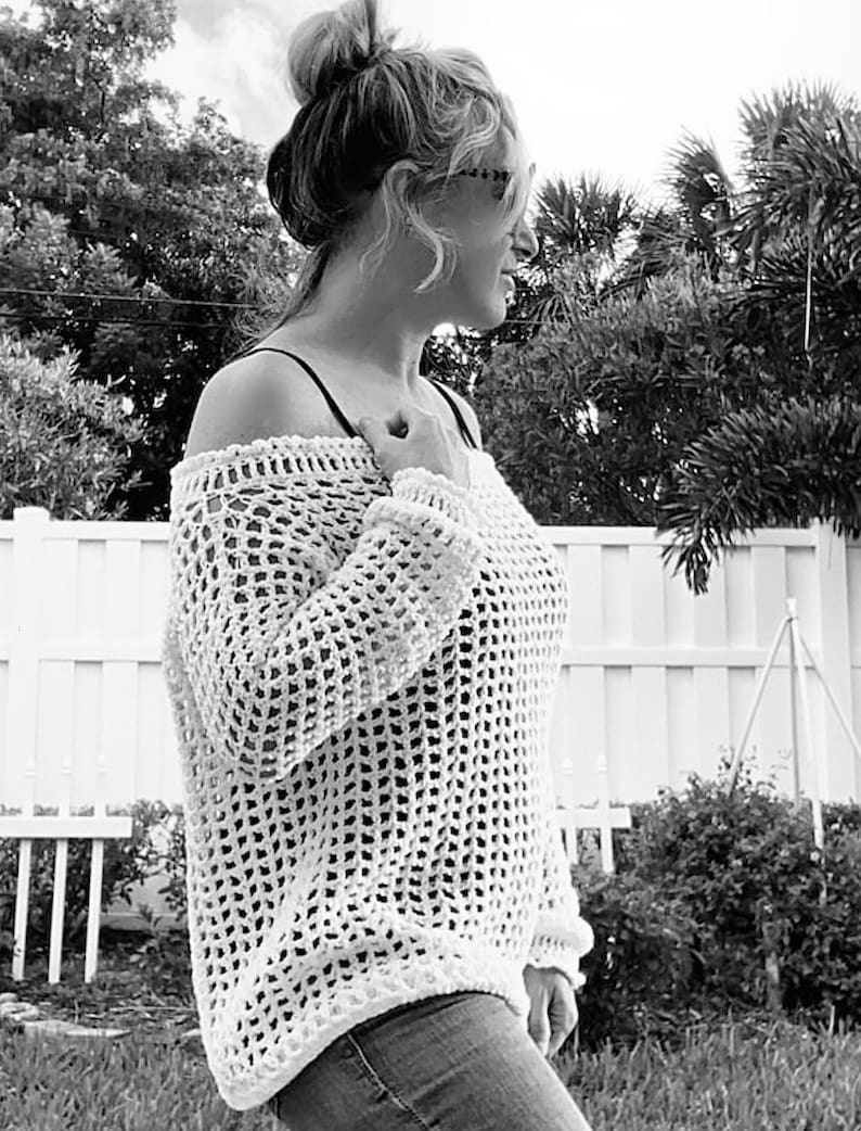 May include: A white, open-weave crochet sweater with long sleeves. The off-the-shoulder design features a loose, airy knit. The sweater is paired with blue jeans. The background includes a white fence and lush greenery.