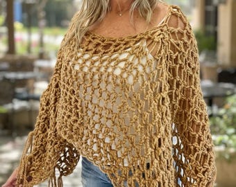 Könnte beinhalten: Ein beigefarbener Häkelponcho mit Fransen. Das offene Design des Ponchos ist sichtbar, und er wird über einem weißen Oberteil und einer blauen Jeans getragen. Der Poncho hat eine lockere Passform und einen Bohemian-Stil.
