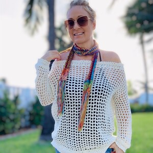 May include: Off-the-shoulder, cream-colored crochet sweater with long sleeves. A colorful, fringed scarf is draped around the neck. The person is wearing sunglasses and blue jeans. The sweater has a loose, open weave.