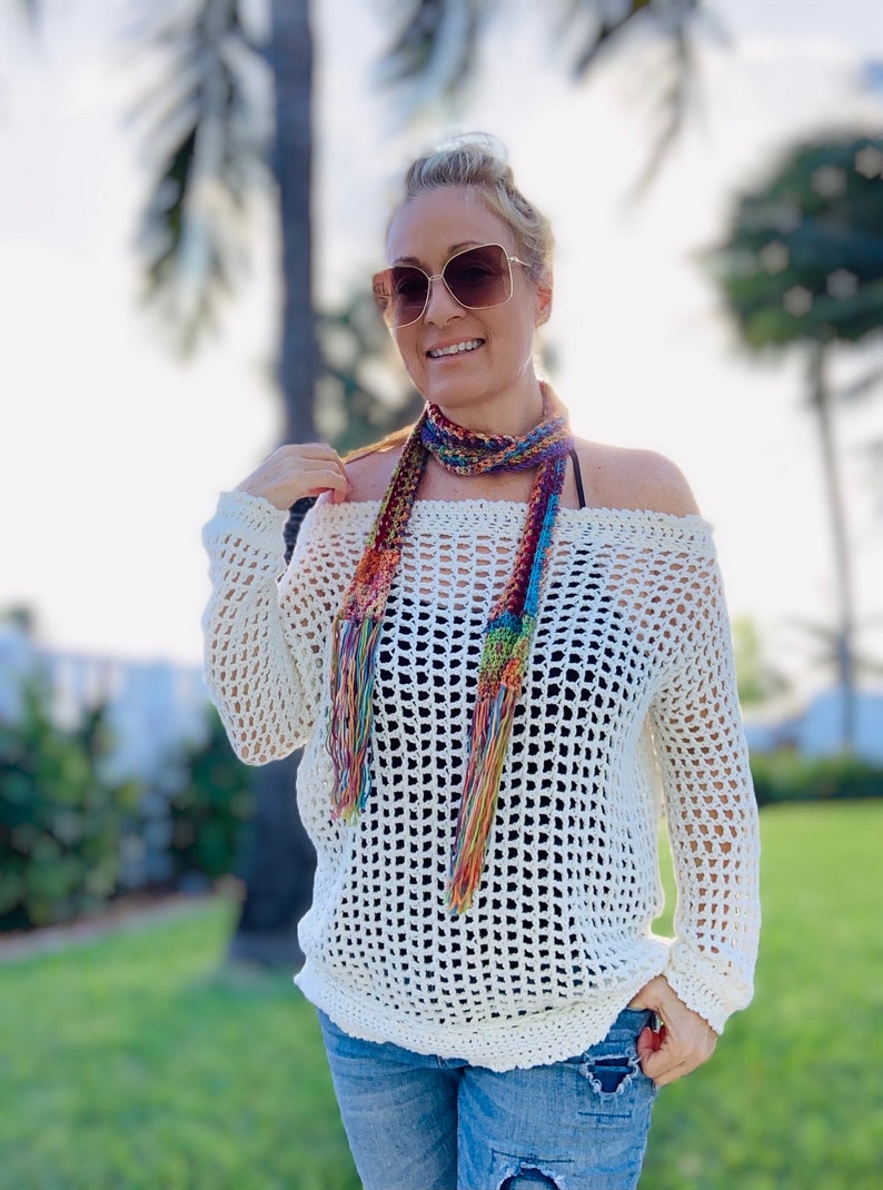 May include: Off-the-shoulder, cream-colored crochet sweater with long sleeves. A colorful, fringed scarf is draped around the neck. The person is wearing sunglasses and blue jeans. The sweater has a loose, open weave.