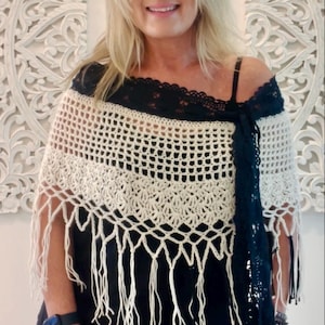 May include: A white crocheted shawl with fringe, worn over a black top with a lace trim. The shawl has a black ribbon tied around the neck.