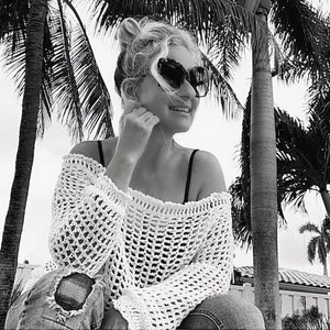 May include: A black and white image of a person wearing a white crochet top, black sunglasses, and ripped jeans. The person is smiling and sitting outdoors with palm trees in the background.
