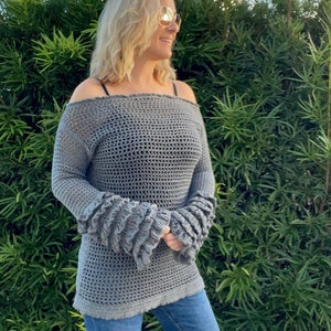 Op de afbeelding: Een grijze, off-the-shoulder gehaakte top met lange mouwen en gerimpelde manchetten. De top wordt gecombineerd met een blauwe jeans. Het haakpatroon is open, waardoor een textuur ontstaat. De mouwen hebben een uniek, gelaagd ruchedesign.