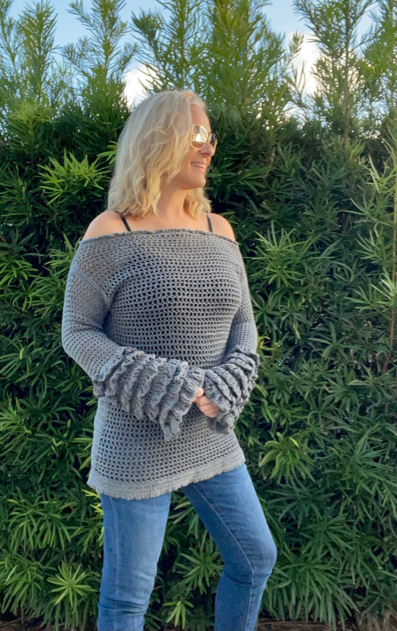 Op de afbeelding: Een grijze, off-the-shoulder gehaakte top met lange mouwen en gerimpelde manchetten. De top wordt gecombineerd met een blauwe jeans. Het haakpatroon is open, waardoor een textuur ontstaat. De mouwen hebben een uniek, gelaagd ruchedesign.