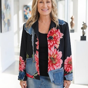 May include: Black blazer with large pink floral print and denim accents. The jacket features a black collar and denim pockets. The outfit includes a black top and a denim mini-skirt. The woman is smiling.