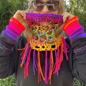 Op de afbeelding: Een kleurrijke gehaakte nekwarmer met franjes. De nekwarmer is gemaakt van verschillende felle kleuren, waaronder roze, paars, oranje en geel. De nekwarmer wordt gedragen door een persoon met een zonnebril.