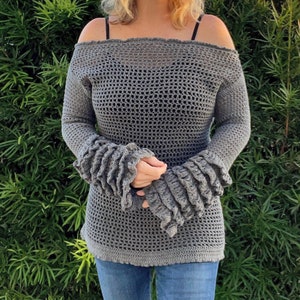 Op de afbeelding: Een grijze, off-the-shoulder gehaakte top met lange mouwen en gerimpelde manchetten. De top wordt gecombineerd met een blauwe jeans. De open structuur van het haakwerk cre&euml;ert een textuur. De mouwen hebben een opvallend, gelaagd ruche-ontwerp.