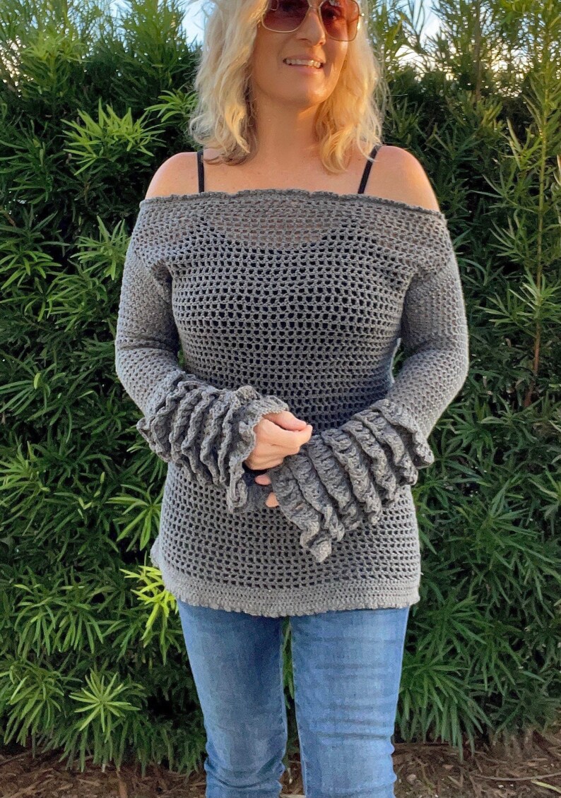 Op de afbeelding: Een grijze, off-the-shoulder gehaakte top met lange mouwen en gerimpelde manchetten. De top wordt gecombineerd met een blauwe jeans. De open structuur van het haakwerk cre&euml;ert een textuur. De mouwen hebben een opvallend, gelaagd ruche-ontwerp.