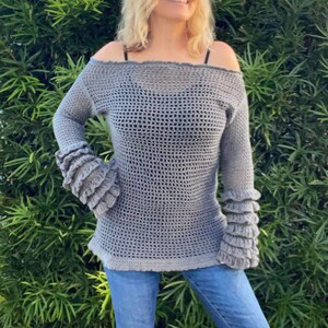 Op de afbeelding: Een grijze, off-the-shoulder gehaakte trui met lange mouwen en gerimpelde manchetten. De trui heeft een losse, open weefsel en een licht uitlopende zoom. Het model draagt een blauwe jeans.
