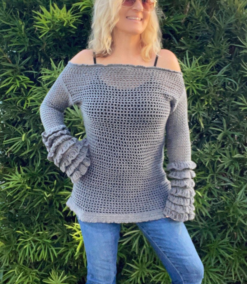 Op de afbeelding: Een grijze, off-the-shoulder gehaakte trui met lange mouwen en gerimpelde manchetten. De trui heeft een losse, open weefsel en een licht uitlopende zoom. Het model draagt een blauwe jeans.