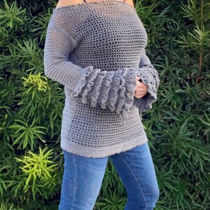 Op de afbeelding: Grijs gehaakt off-the-shoulder top met lange mouwen en uitlopende manchetten. De top wordt gecombineerd met een blauwe jeans. De open weving van het haakwerk cre&euml;ert een lichte uitstraling. De mouwen hebben een gerimpeld ontwerp.