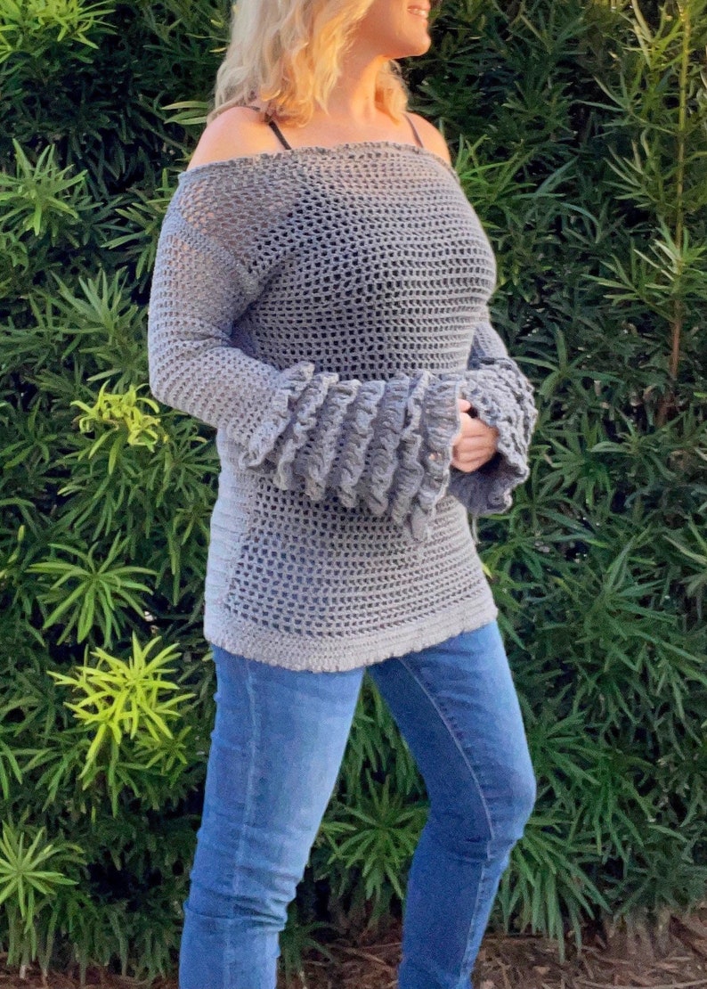 Op de afbeelding: Grijs gehaakt off-the-shoulder top met lange mouwen en uitlopende manchetten. De top wordt gecombineerd met een blauwe jeans. De open weving van het haakwerk cre&euml;ert een lichte uitstraling. De mouwen hebben een gerimpeld ontwerp.