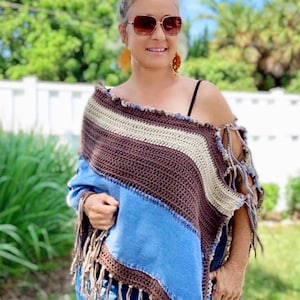 Op de afbeelding: Een bruin, beige en blauw gebreid poncho met franjes. De poncho wordt gedragen over een zwart tanktop en een blauwe jeans.