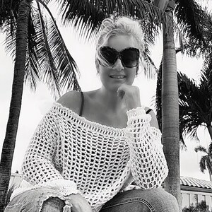 May include: A black and white image of a person wearing sunglasses and a white, open-weave crochet sweater. The sweater has long sleeves and an off-the-shoulder design. The background features palm trees.