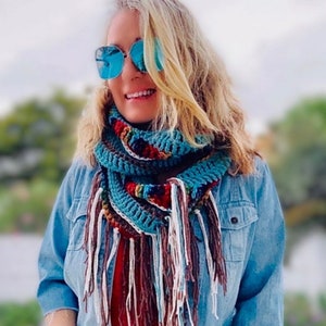 Peut inclure: Une femme portant une veste en jean bleue et une écharpe infinie en crochet avec des rayures colorées et de longues franges. L'écharpe est faite de plusieurs couleurs, dont le bleu, le vert, le rouge et le blanc.